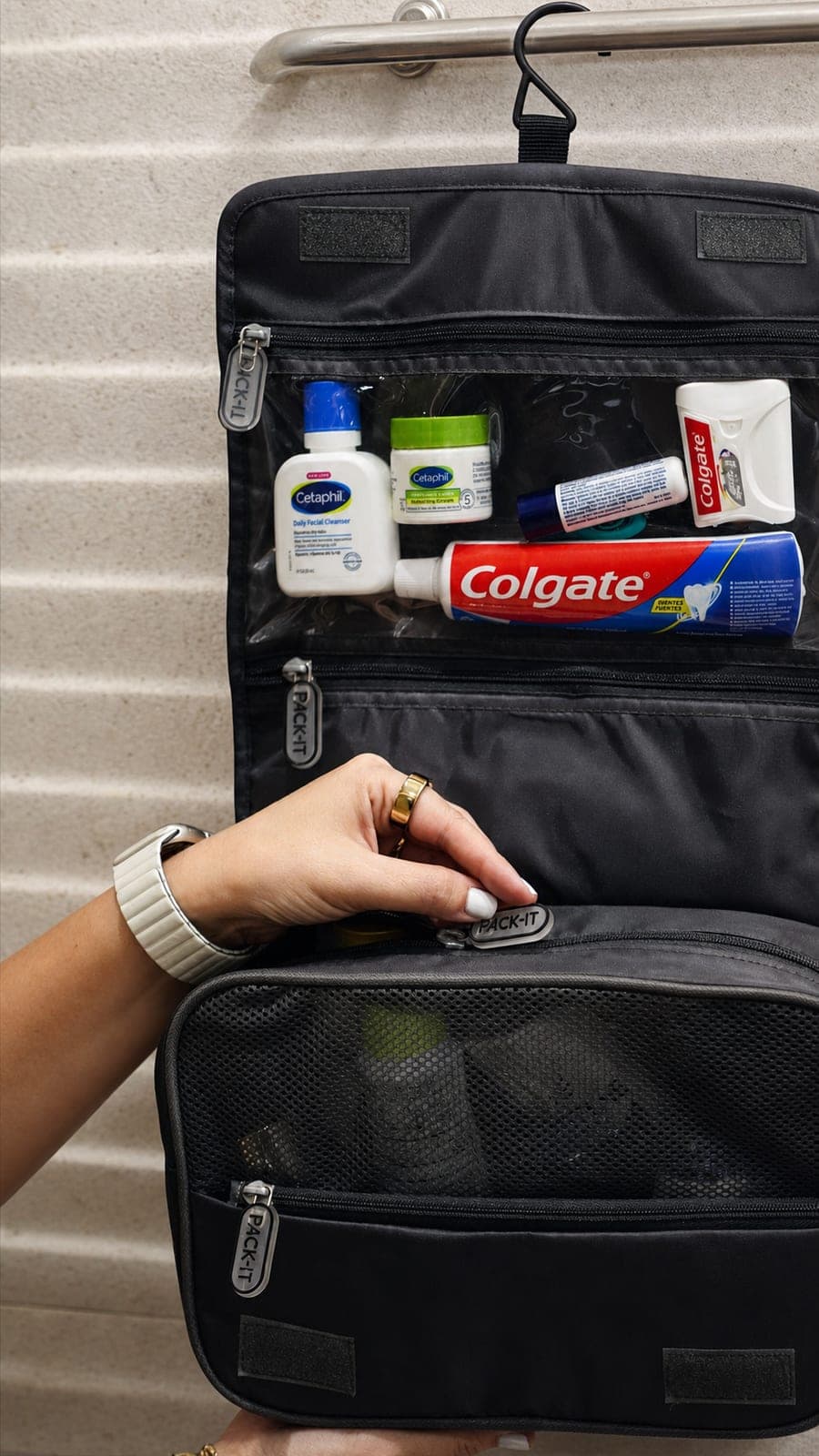 Neceser Smart Cube gris colgado de una barra de baño en un hotel, abierto con bolsillo transparente que muestra Cetaphil y Colgate,…