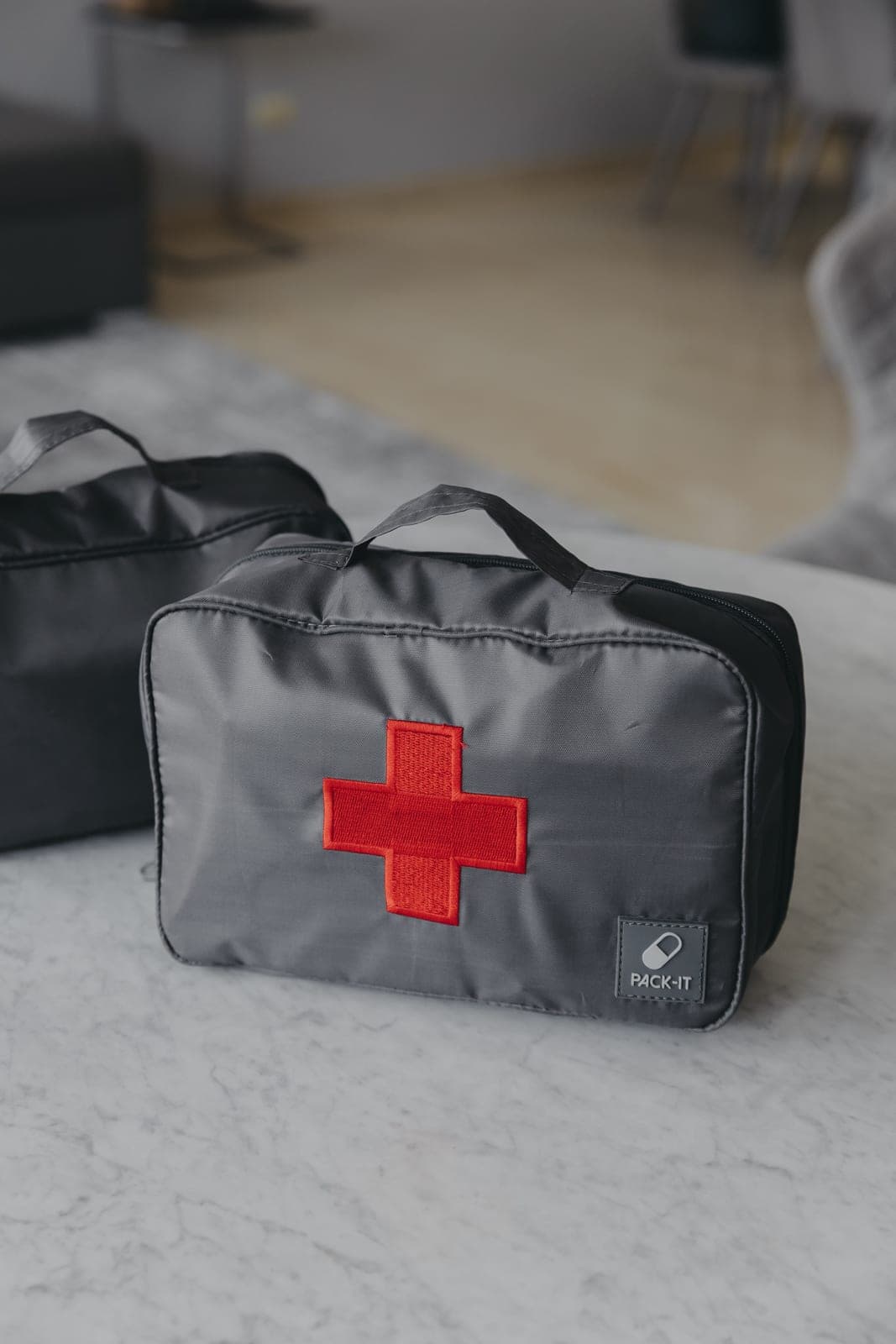 Emergency Cube gris con cruz roja bordada y etiqueta PACK-IT sobre una mesa de mármol, con otro estuche oscuro desenfocado al fondo en…